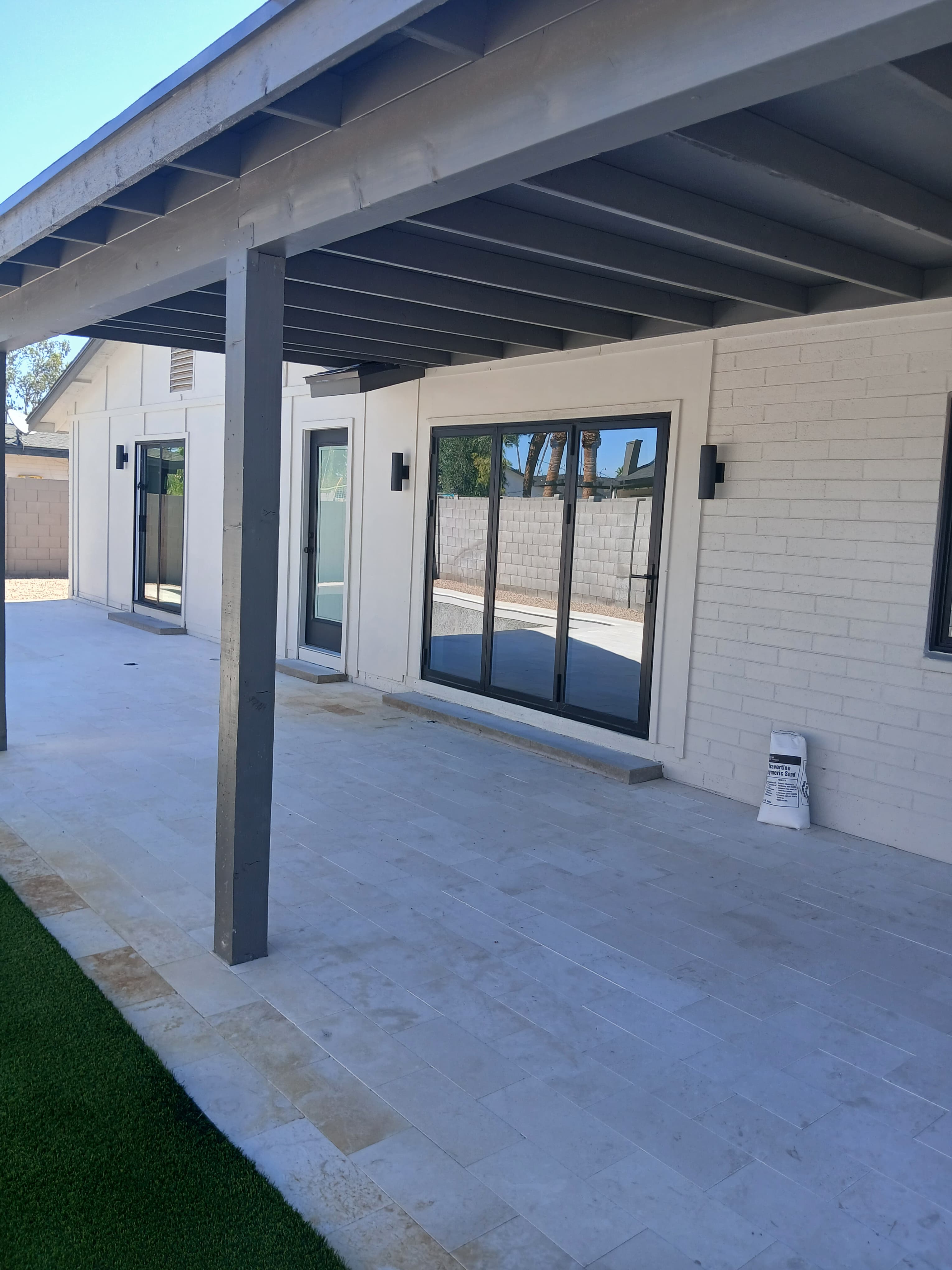 White brick house with black-framed glass doors, a grey pergola, and a light stone patio.
