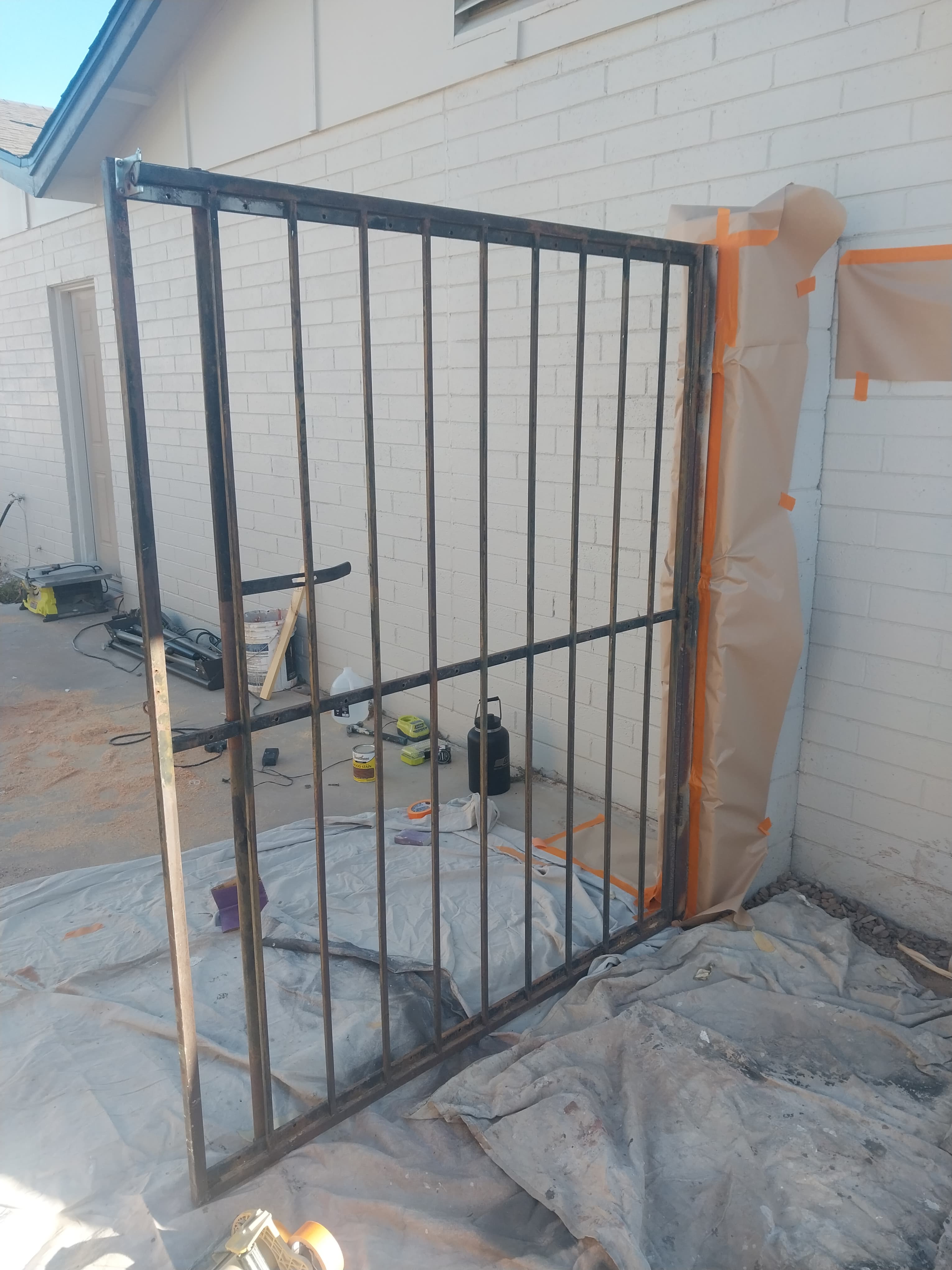 Metal gate frame on drop cloths against a white brick wall being prepared for painting.